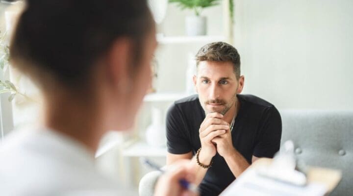 Man sitting across from a therapist during a counseling session, listening intently with his hands clasped under his chin.