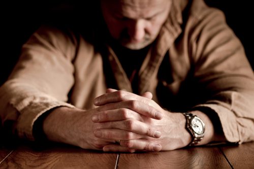 older man sitting at table with hands clasped and head down - men