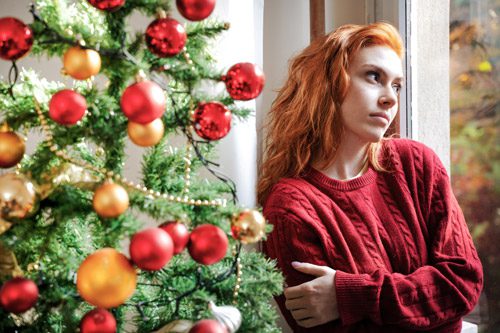 pretty young red haired woman looking out the window while standing next to Christmas tree - looking lonely - loneliness