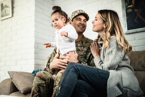 handsome man in military fatigues holding toddler daughter on couch with smiling wife - veteran