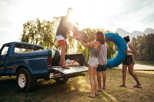 group of young adult friends unloading items such as an inflatable inner tube and a picnic basket from a pickup truck parked near lake or river - staying sober