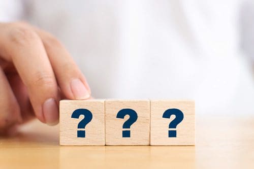 three wooden blocks with navy blue question marks on them lined up by a man on wooden table - he is out of focus and cropped out of image - intervention