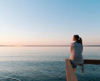 Woman sitting peacefully on a wooden railing overlooking calm water at sunset, reflecting quietly in a serene, open landscape