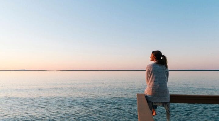 Woman sitting peacefully on a wooden railing overlooking calm water at sunset, reflecting quietly in a serene, open landscape