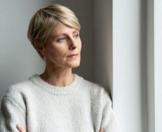 Woman standing by a window with a thoughtful expression, suggesting reflection, concern, or emotional contemplation.