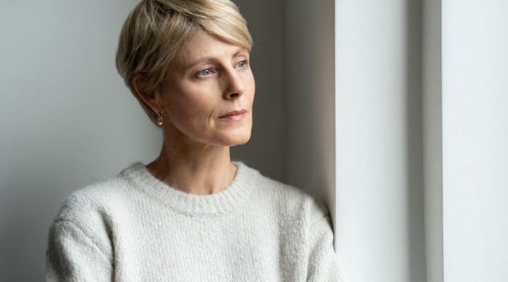 Woman standing by a window with a thoughtful expression, suggesting reflection, concern, or emotional contemplation.