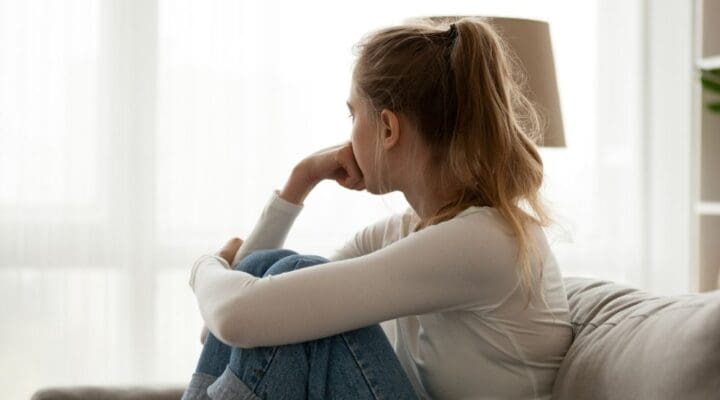 Young woman sitting on a couch with her knees pulled up, gazing out the window with a thoughtful and distant expression.