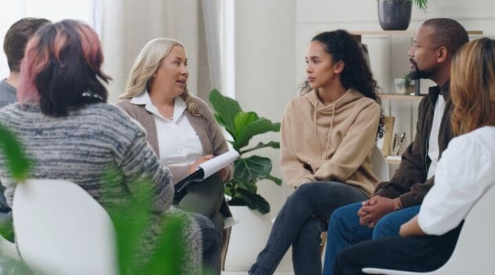 A diverse group of people sit in a circle during a support group meeting. A woman holding a clipboard speaks while others listen attentively. The setting is bright and welcoming, with plants and natural light.