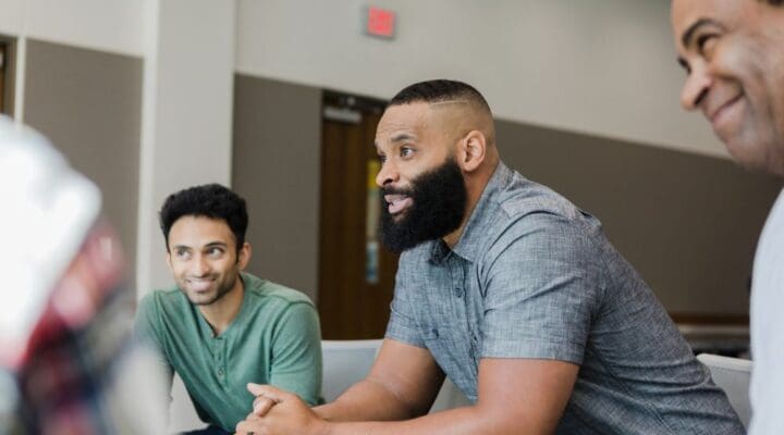Group of diverse men sitting in a support group setting, actively engaged in conversation and smiling.
