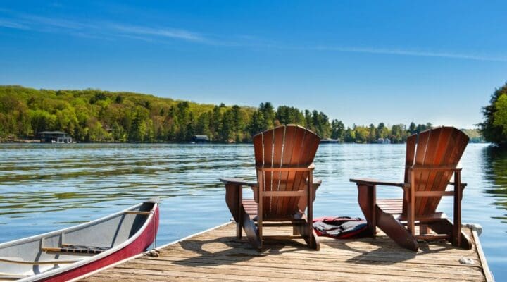 Two Adirondack chairs and a red canoe on a sunny lakeside dock, surrounded by trees and calm water—capturing a peaceful summer day.