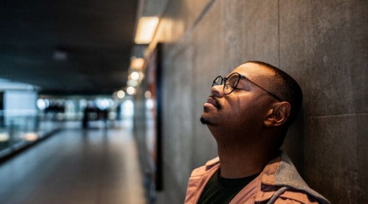 A man wearing glasses leans against a wall in a dimly lit corridor, eyes closed and looking distressed, symbolizing anxiety and emotional struggle.