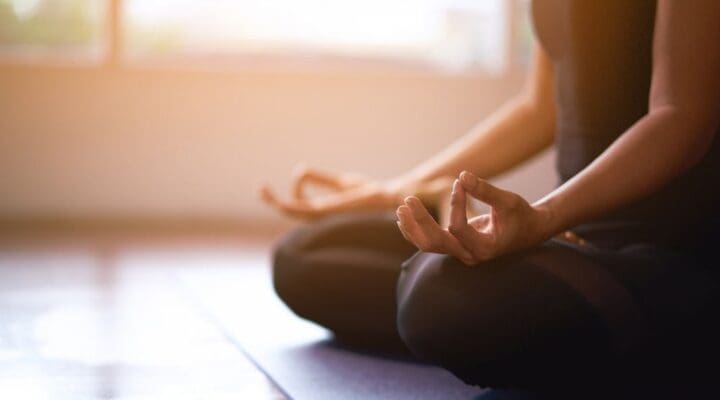 A person sitting cross-legged on a yoga mat indoors, with hands resting on knees in a meditative pose. Soft sunlight filters through large windows in the background, creating a peaceful and serene atmosphere.