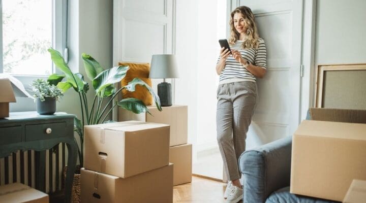 Woman leaning against a wall and looking at her phone, surrounded by moving boxes in a bright living room, symbolizing life transitions.