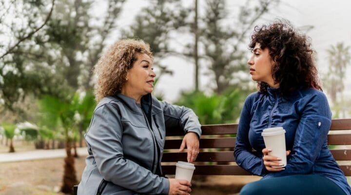Two women sit on a park bench holding coffee cups, engaged in a serious conversation, symbolizing support, boundaries, and connection.