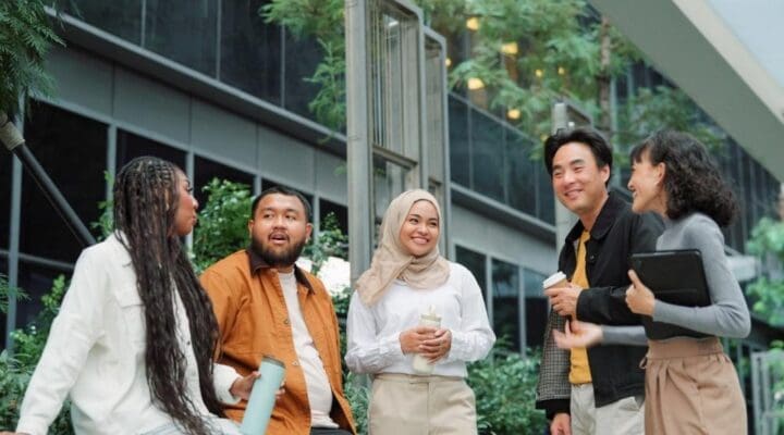 Group of coworkers standing and chatting outside an office building, smiling and holding coffee cups and water bottles in a relaxed atmosphere.