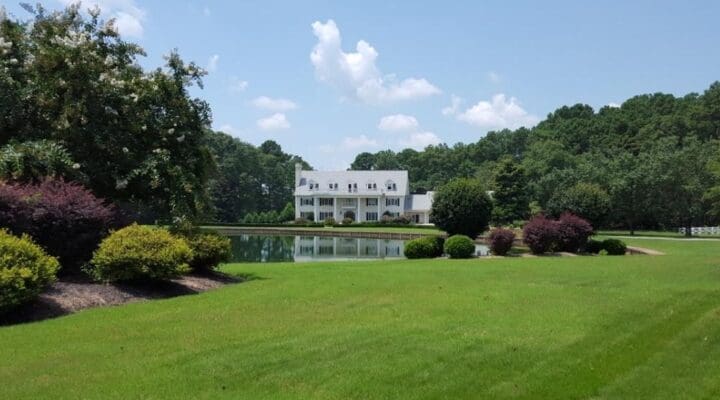 A large white house with columns stands in front of a pond, surrounded by green lawns, trees, and landscaped bushes on a sunny day.