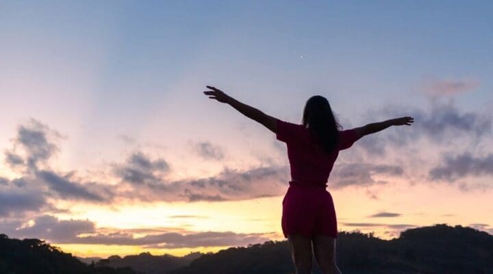 A woman stands outdoors at sunrise with her arms outstretched, facing the horizon. She appears peaceful and empowered as the sky glows with soft pink and purple tones.