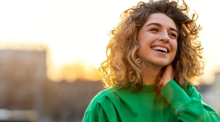 A smiling woman with curly hair standing outdoors at sunset, looking joyful and relaxed.