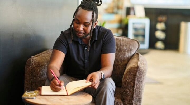 A man sits in a comfortable chair writing in a notebook, journaling thoughtfully in a relaxed indoor setting.