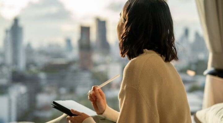 A person sits by a window overlooking a city, holding a notebook and pen while reflecting quietly in a calm, sunlit room.