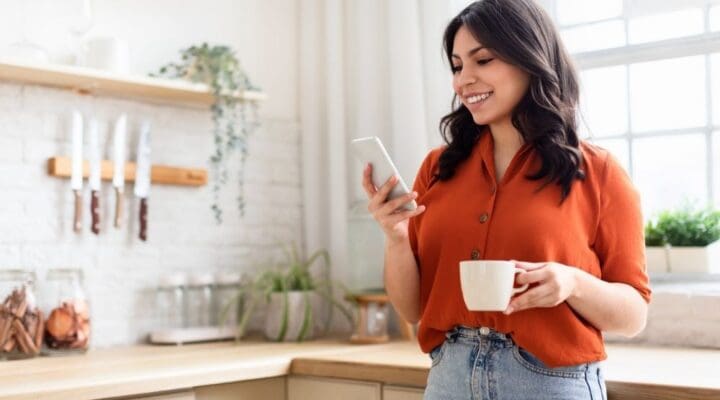 Smiling young woman enjoying her coffee while using her smartphone in a homey kitchen setting