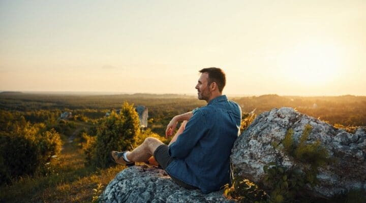 Person sitting on a rocky overlook at sunset, reflecting quietly while overlooking a wide, peaceful natural landscape.